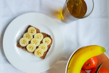 assiette contenant une tartine de morceaux de banane sur une couche de pâte à tartiner à l'amande avec un bol rempli de fruit et un verre de thé